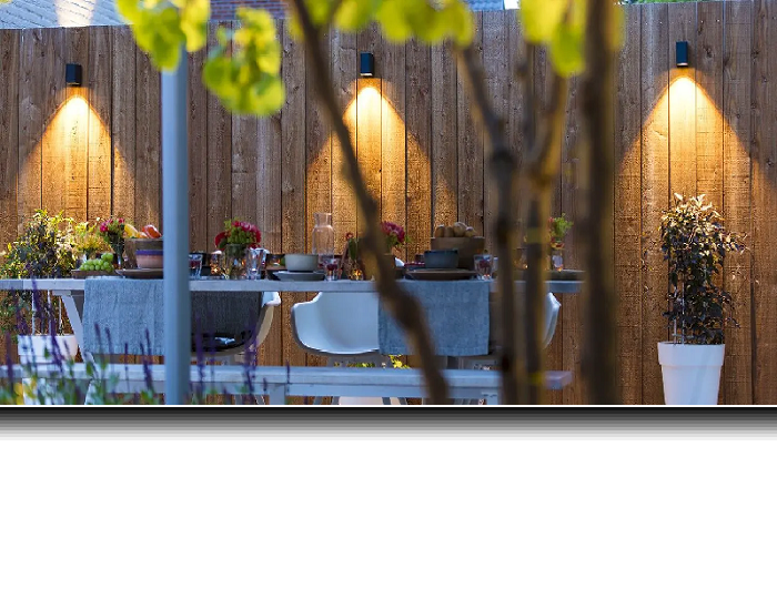 Een terras met grijze terrastegels met daarop een houten tuintafel met vier met vier grijze stoelen. Op de tafel vier borden, bestek een bloemenvaas met rode bloemen en twee folders. 
